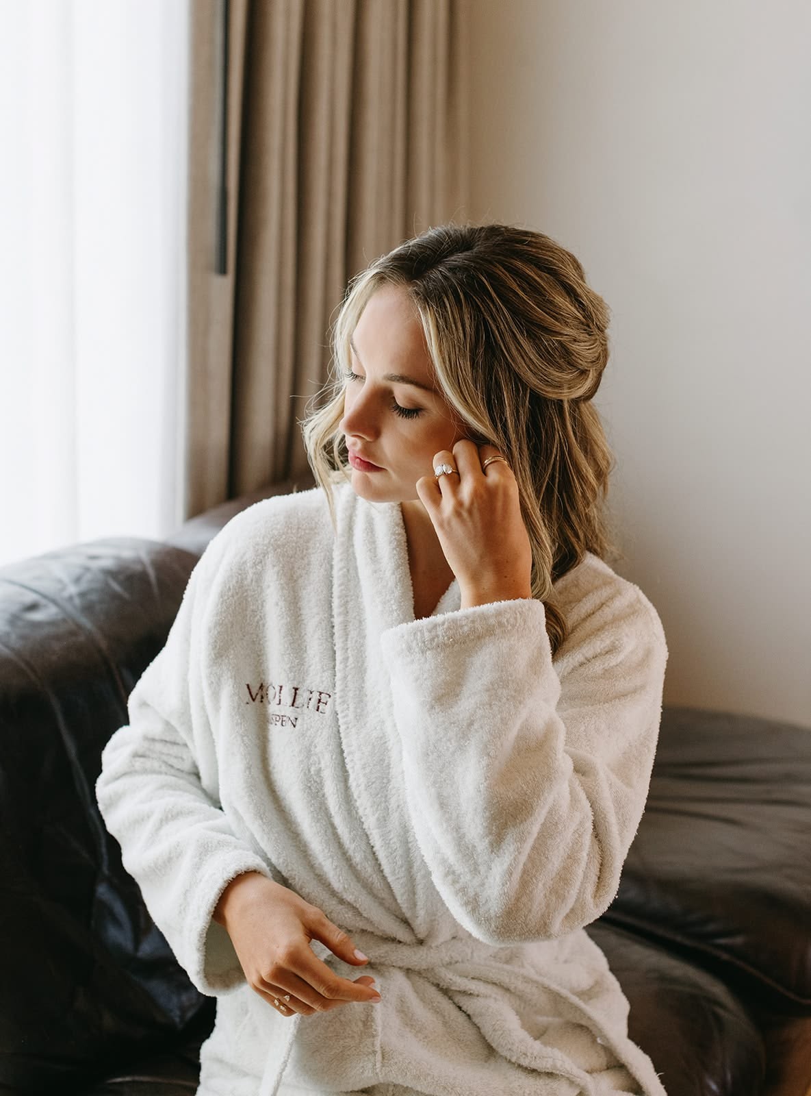 Bride getting ready in a Mollie Aspen robe, putting on earrings by the window