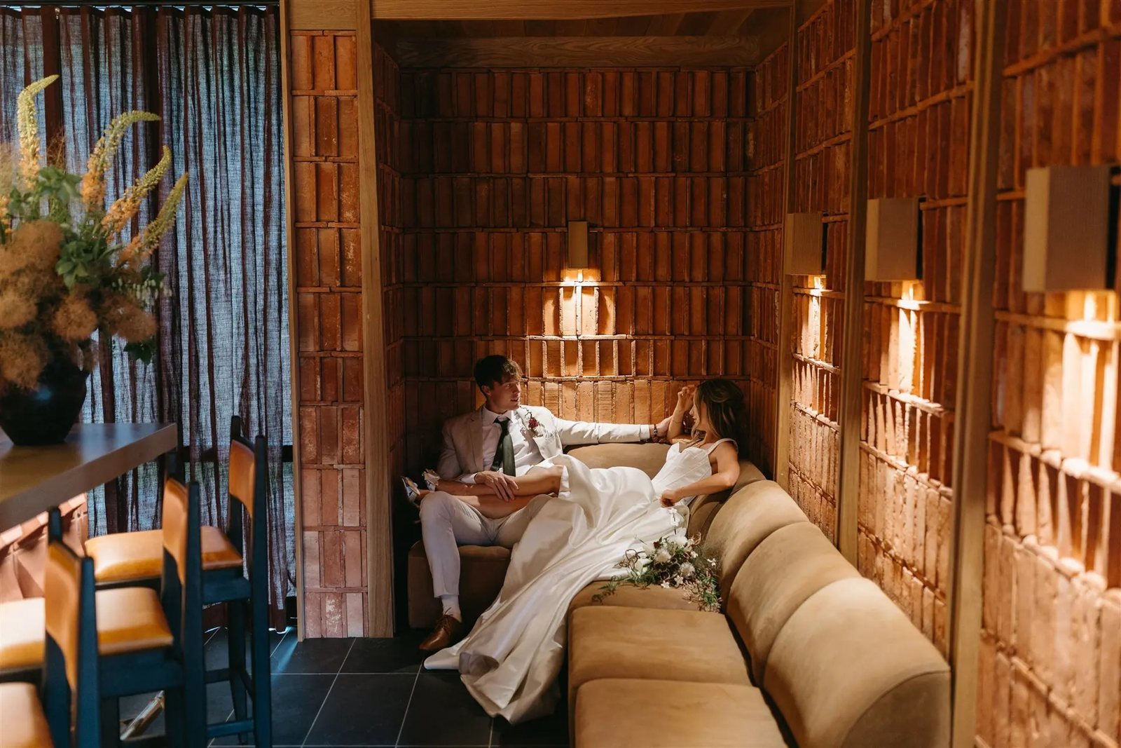 Bride and groom relaxing on a velvet banquette in a warm brick-lined nook