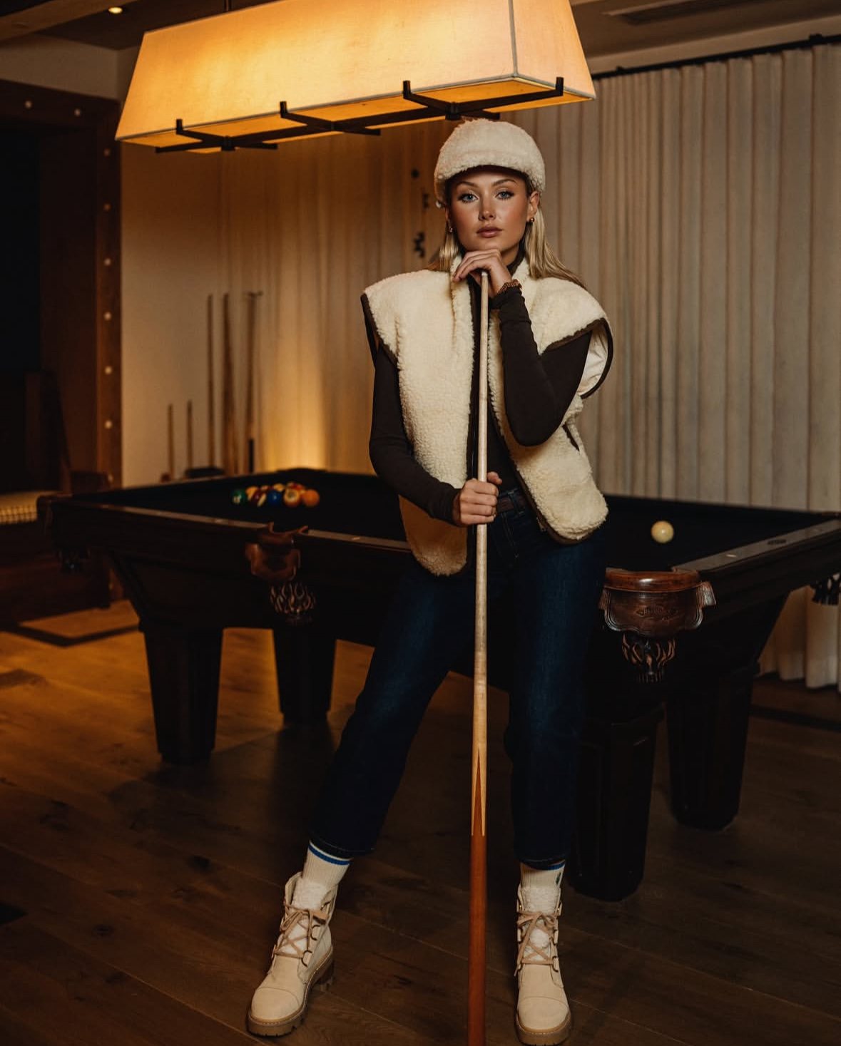 Full-length in a cream shearling cap and vest, dark jeans and cream boots, leaning on a pool cue