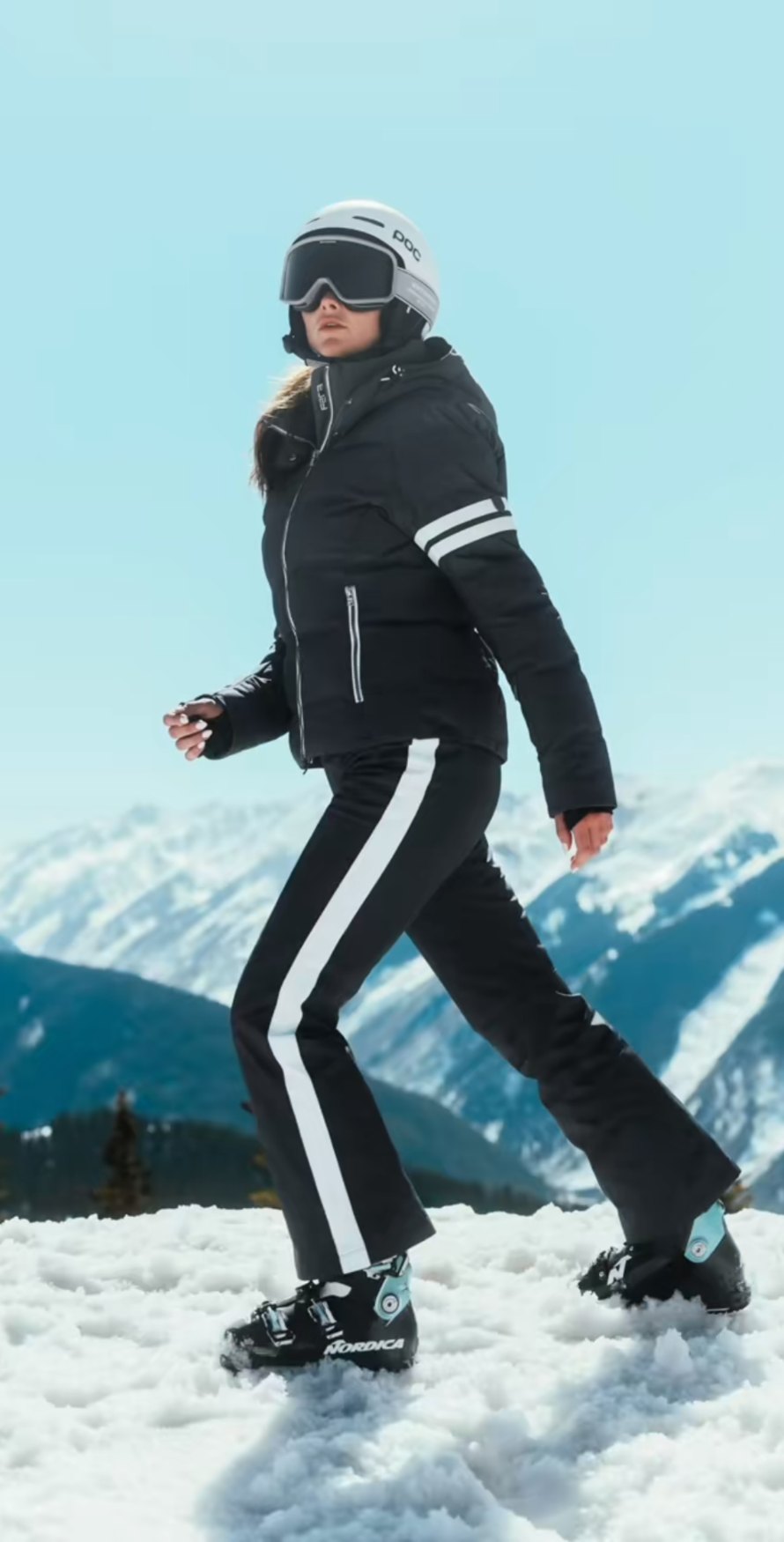 Walking in a black down jacket and striped black and white ski pants with snowy peaks behind