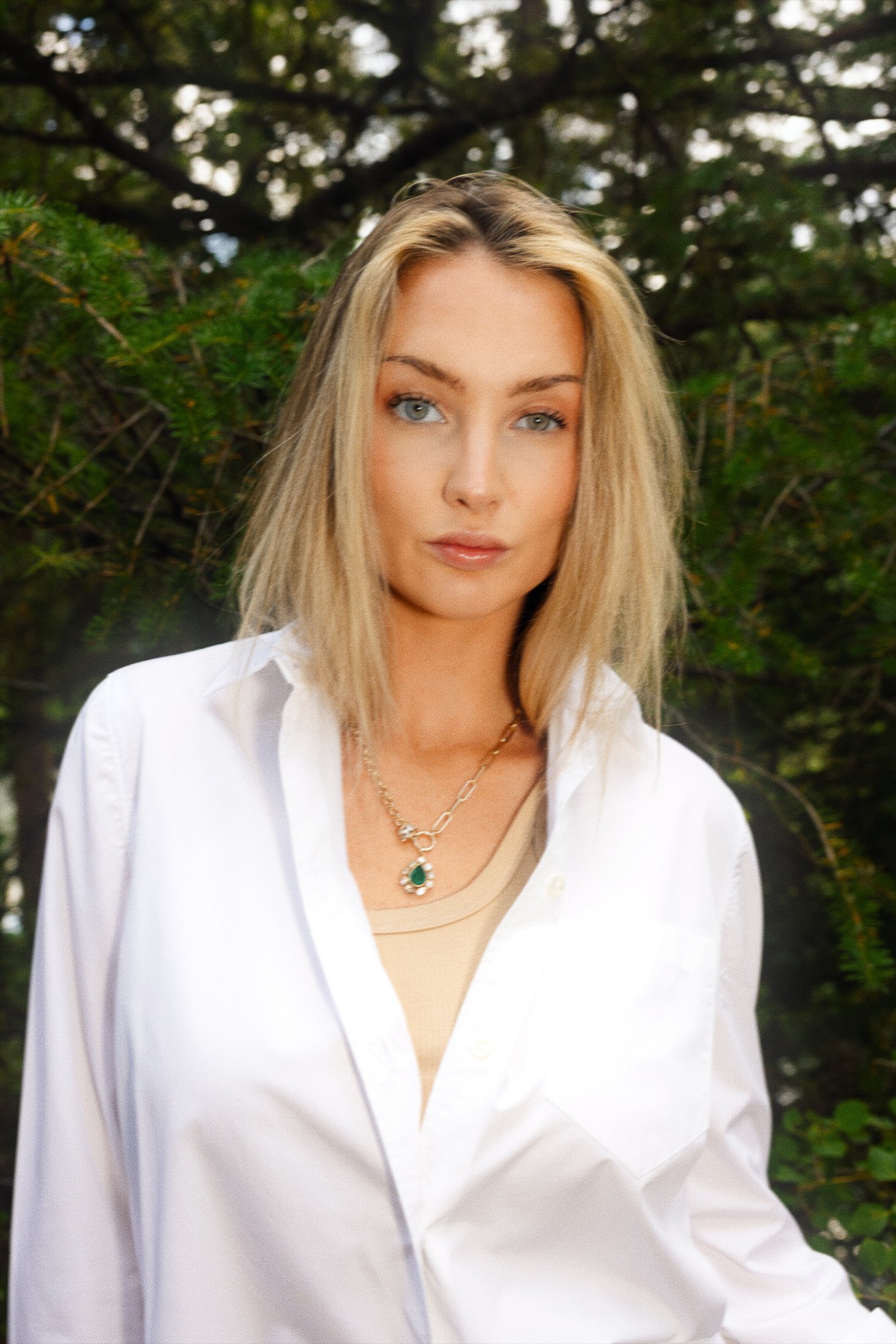 Direct-gaze portrait in an open white shirt with an emerald pendant necklace, pine trees behind