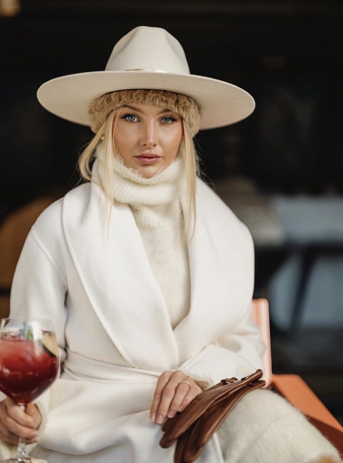 Seated portrait in a cream wide-brim hat, fur headband and white wrap coat, holding a red cocktail and leather gloves