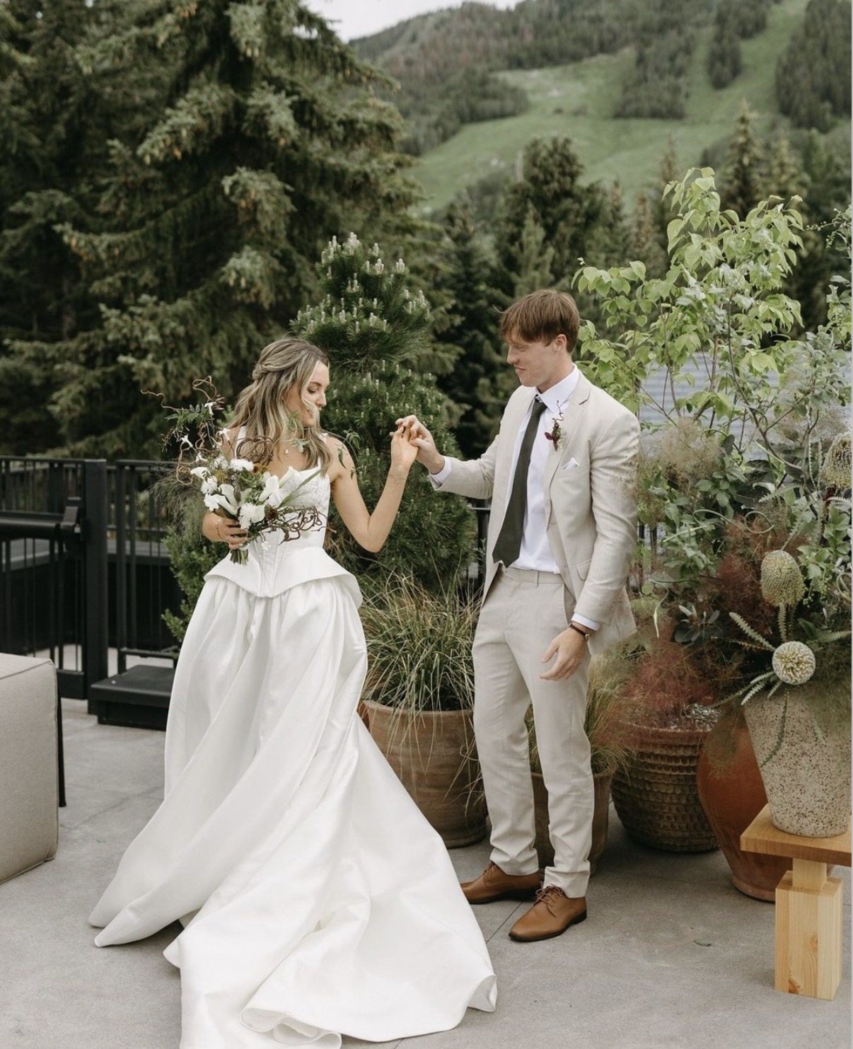 Bride in a satin gown and corset bodice dancing and holding the groom's hand on a terrace with a green mountain behind
