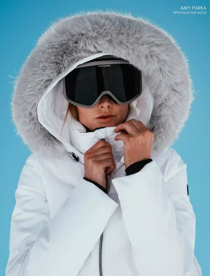 Close-up in a white parka with a grey fur hood and black ski goggles, against a blue sky