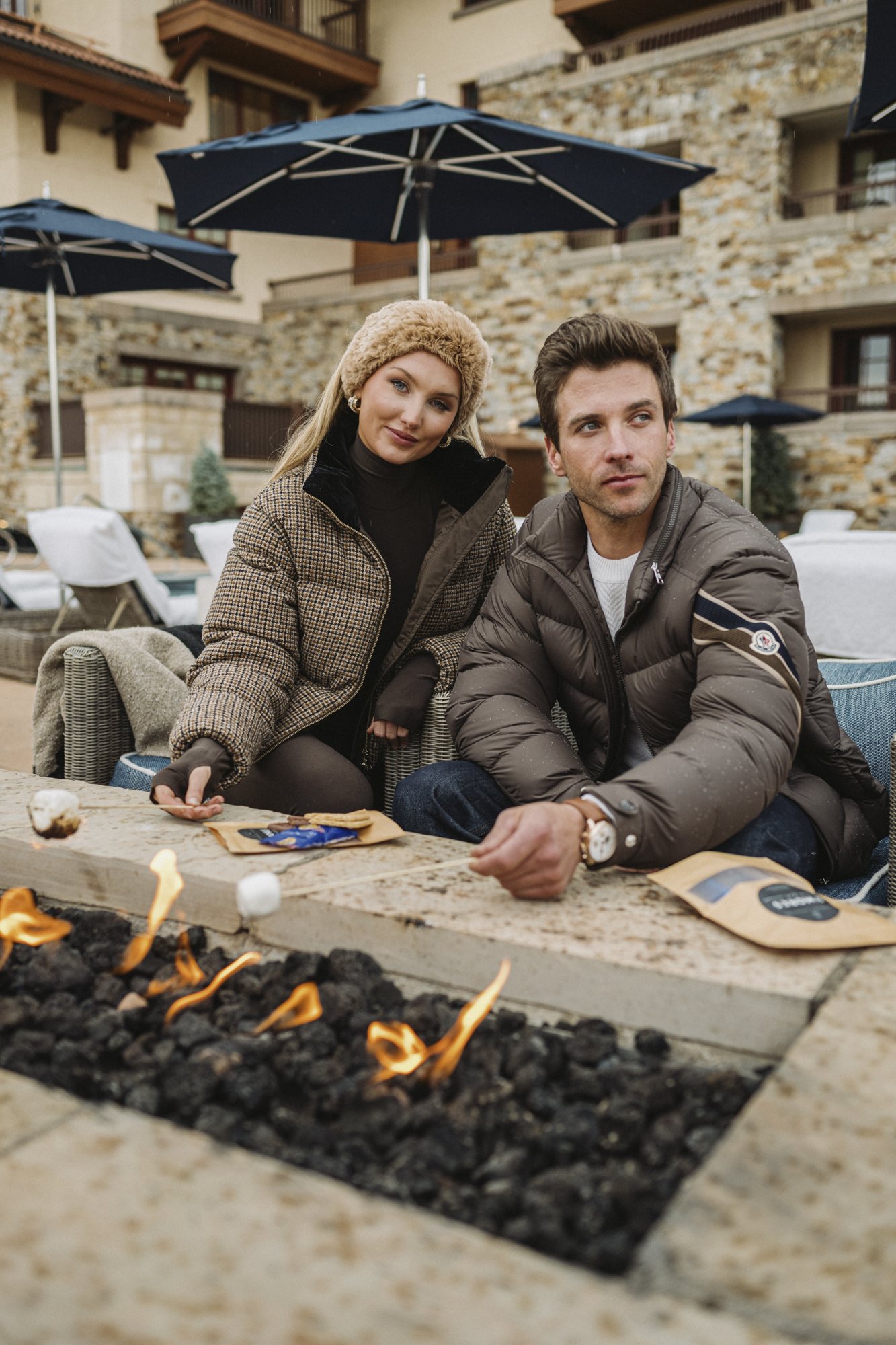 Seated with a male model at an outdoor fire pit, in a fur hat and houndstooth puffer, roasting marshmallows