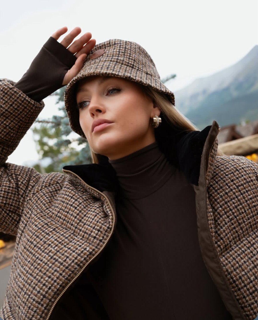 Portrait in a houndstooth bucket hat and puffer jacket over a brown turtleneck, mountains behind