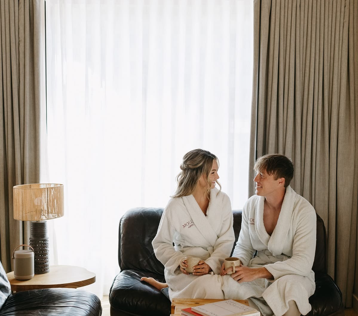 Bride and groom in matching Mollie Aspen robes sharing coffee on a leather sofa by a sunlit window