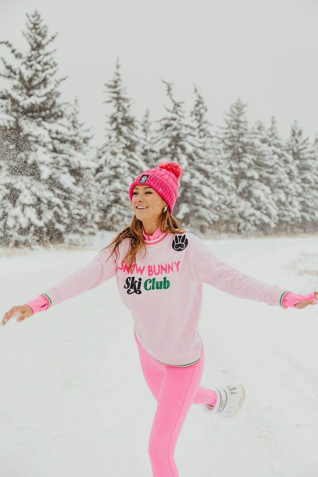 Running and smiling in a pink Snow Bunny Ski Club sweatshirt, pink beanie and pink leggings in falling snow