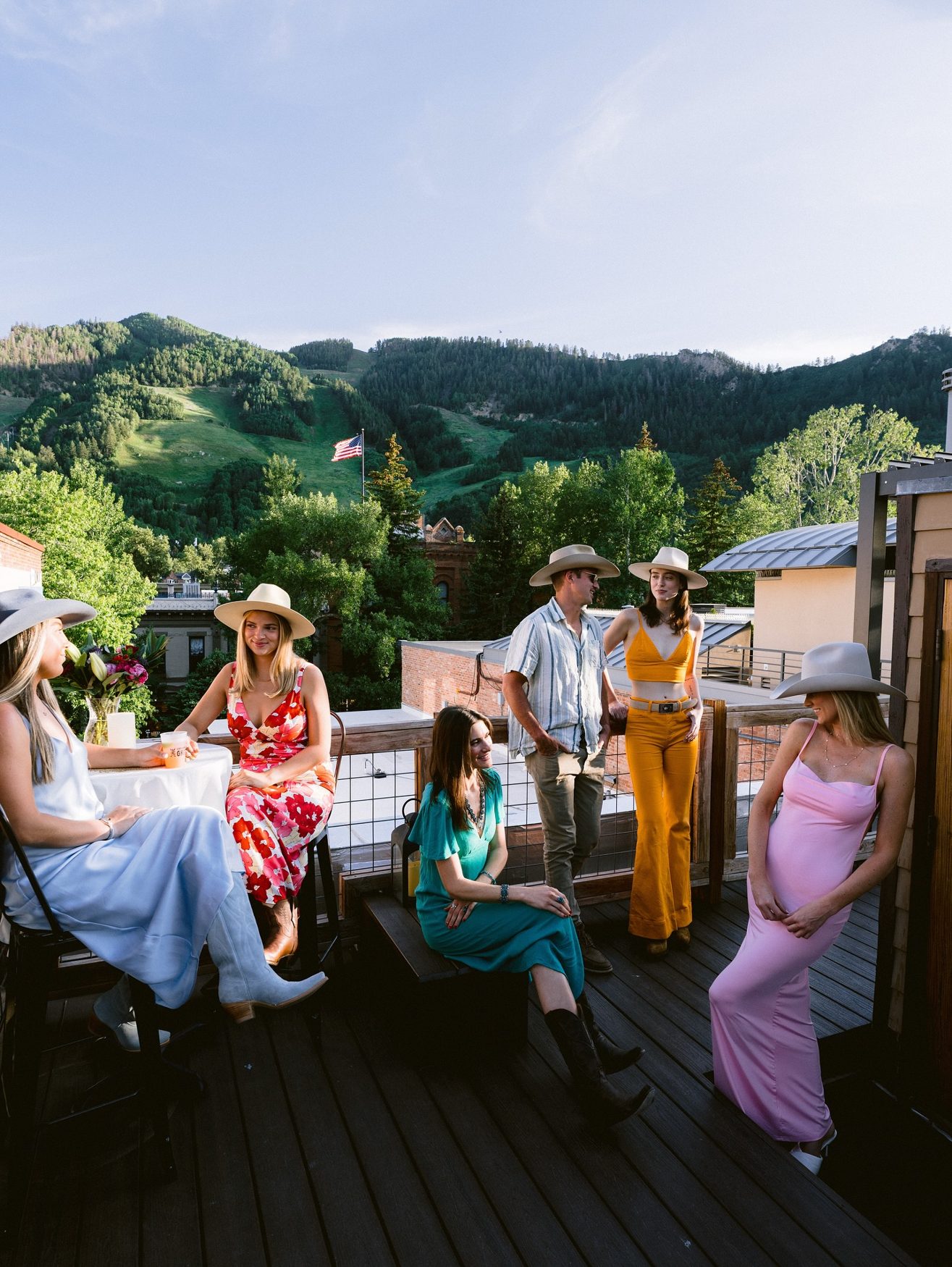 Group in cowboy hats on a mountain-view rooftop at golden hour, model in a pink slip dress at right
