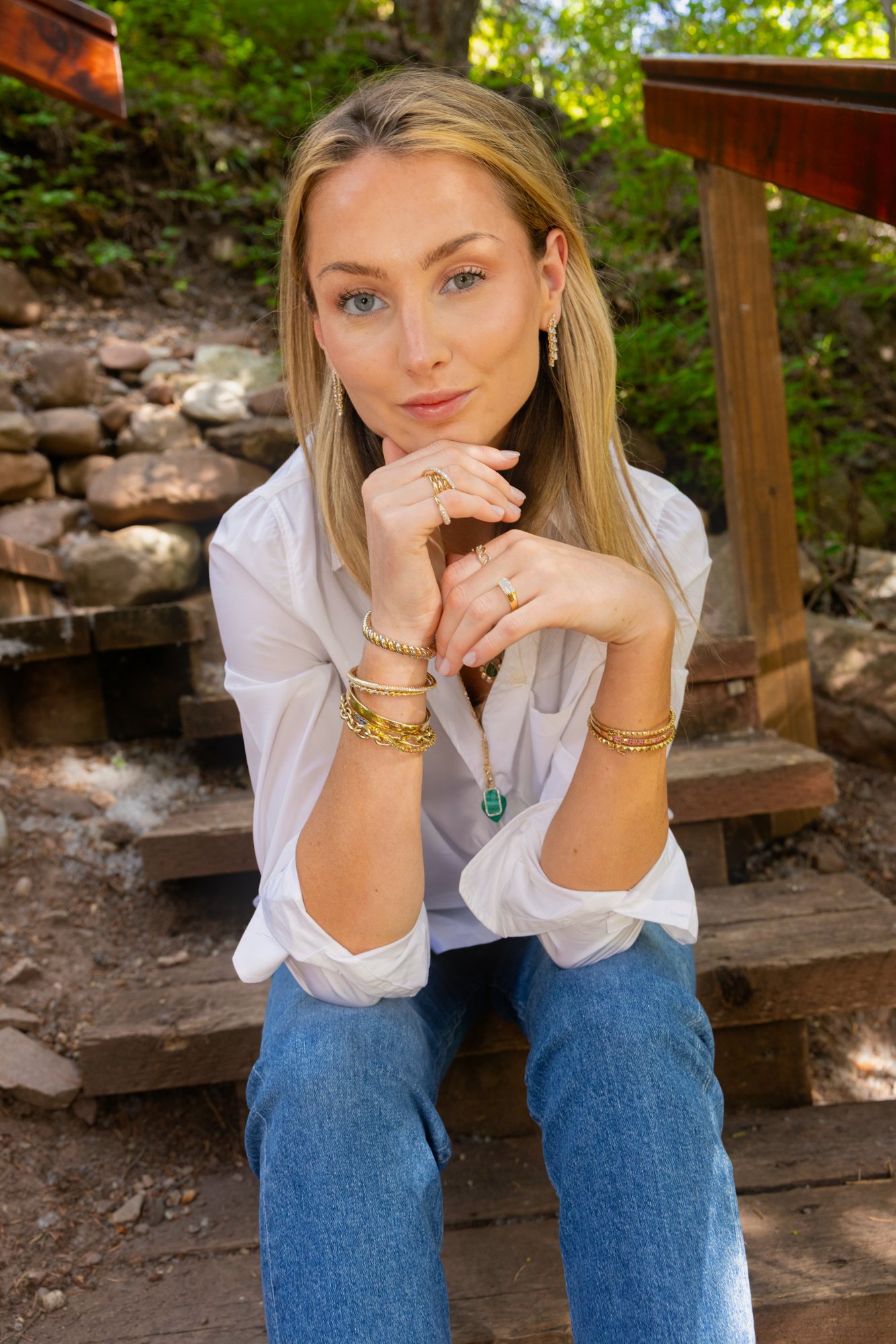 Seated on wooden steps in a white shirt and jeans, chin resting on hands stacked with gold rings and bracelets