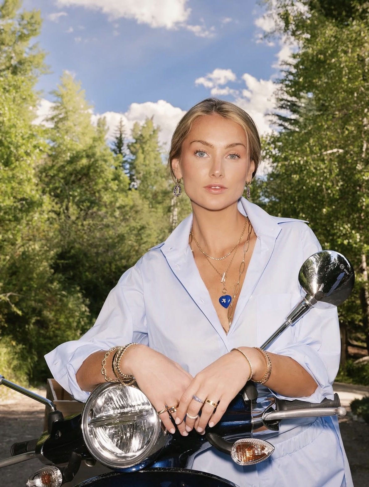 Portrait of Genevieve Brewster in a pale blue shirt with layered gold jewelry, leaning on a scooter in the trees