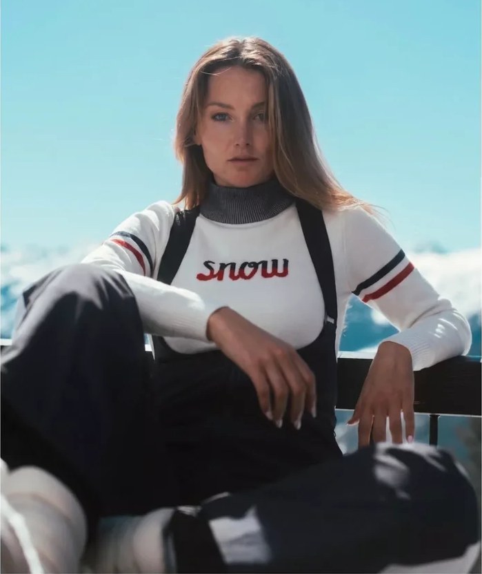 Direct-gaze portrait in a white knit ski sweater with striped sleeves and black ski bibs against a blue sky