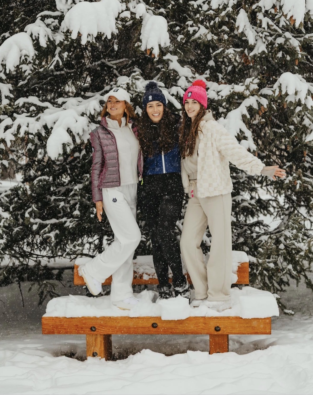 Three friends standing on a snowy bench in front of snow-covered pines, Genevieve at left in a white cap, plum puffer and white pants
