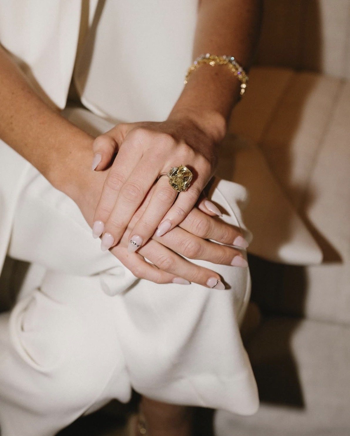 Close-up of crossed hands with a large yellow diamond ring and a gold bracelet, in white tailoring
