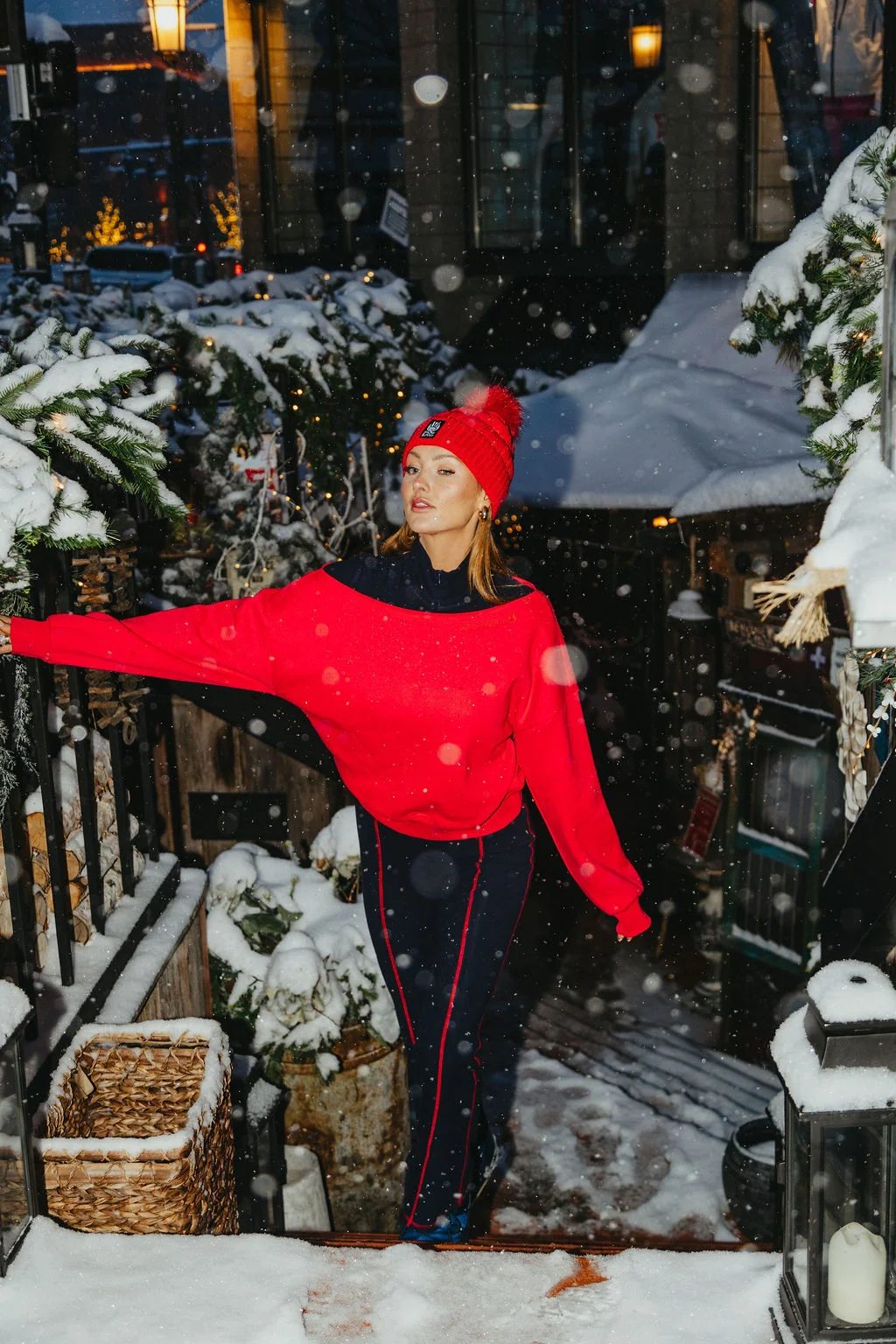 Model in a red pom beanie and red sweatshirt reaching toward a snowy railing at night in falling snow