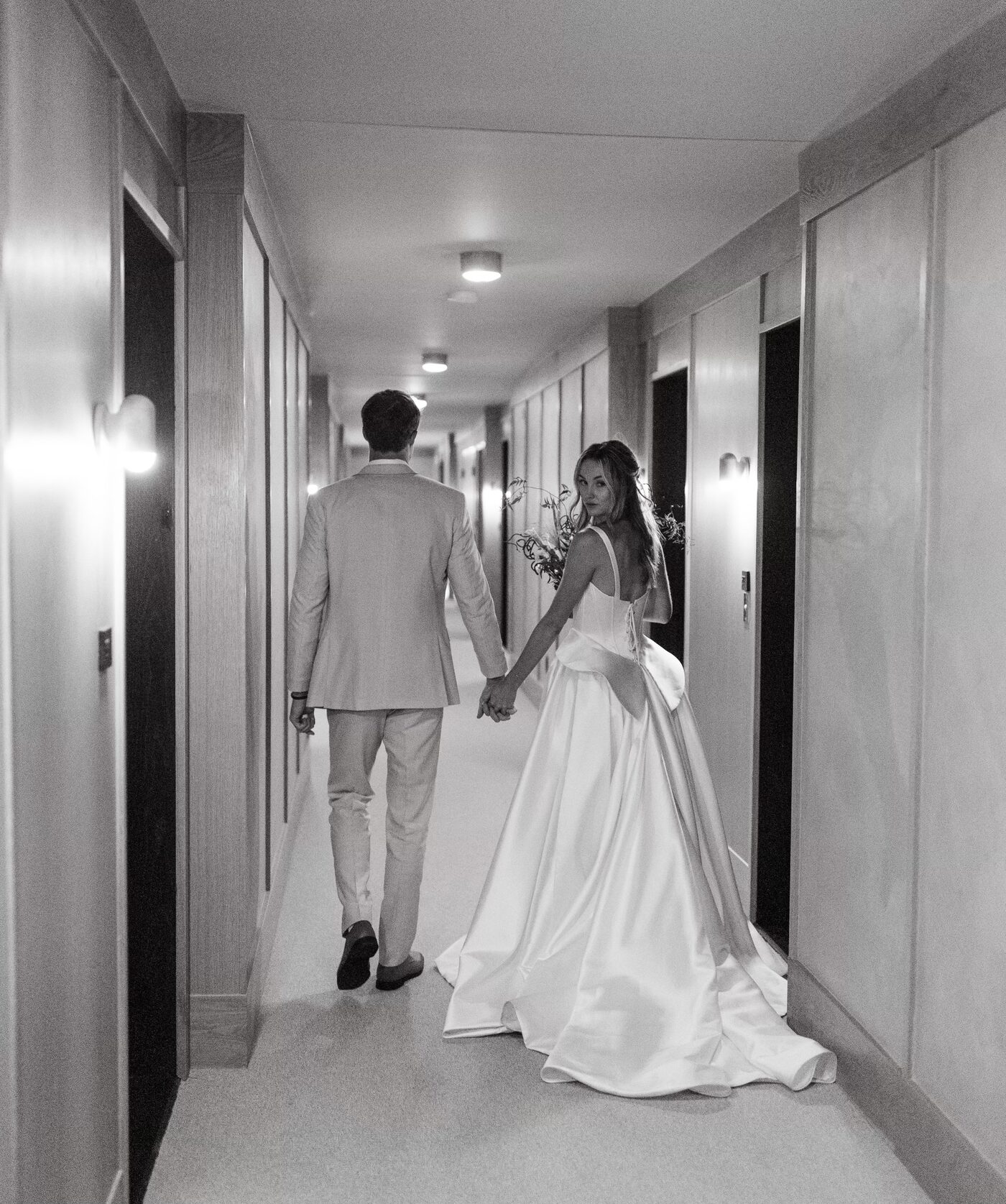 Black and white image of a bride in a satin gown looking back while holding a groom's hand in a hallway