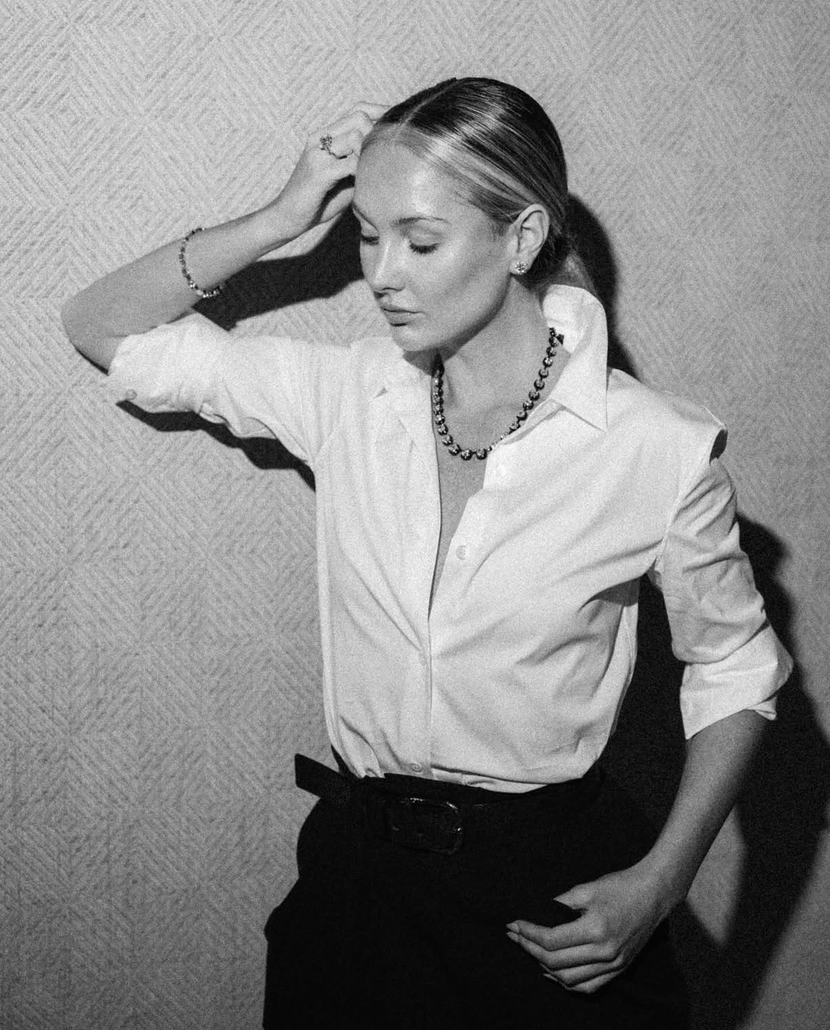 Black and white portrait in a white button-down and black trousers, hand in her hair, against a patterned wall