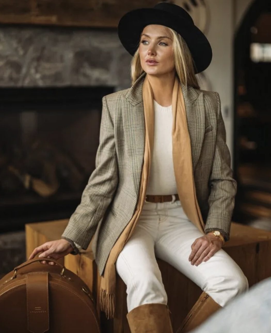 Model in a black hat, plaid blazer and camel scarf by a fireplace