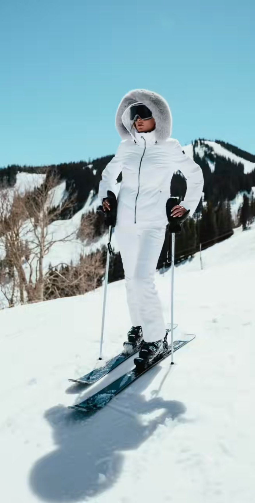 Standing on skis in a white parka with a fur-trimmed hood and white ski pants, mountains behind